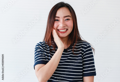 Studio portrait shot of Asian young shy embarrassing female model in casual clothing smiling show teeth look at camera holding raised hand on cheek feeling excited surprised on white background © Bangkok Click Studio