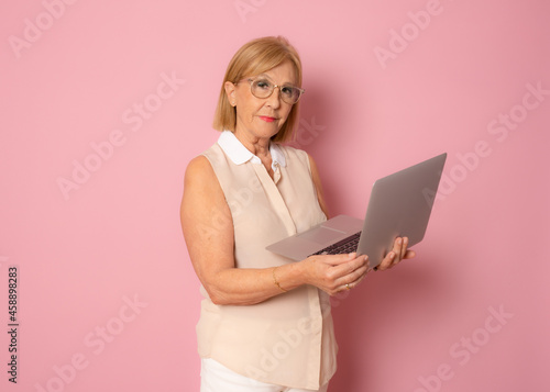 Cheerful mature woman using laptop computer standing isolated over pink background