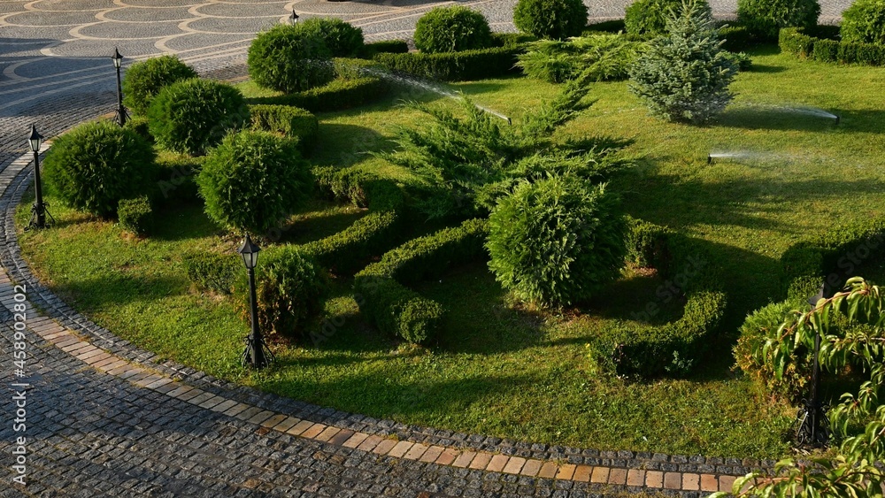 Landscaped garden watering ball shaped mowed shrubs at round lawn with retro lantern streetlights. Ornate garden landscape design in park. Landscaping and gardening