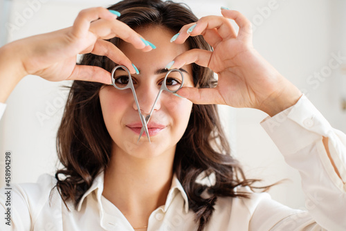 Funny face with nail scissors. Comic young woman. Burnette girl in white holding manicure scissors in front of your eyes just like glasses. Concept of manicure, nail care, beauty Selected focus photo