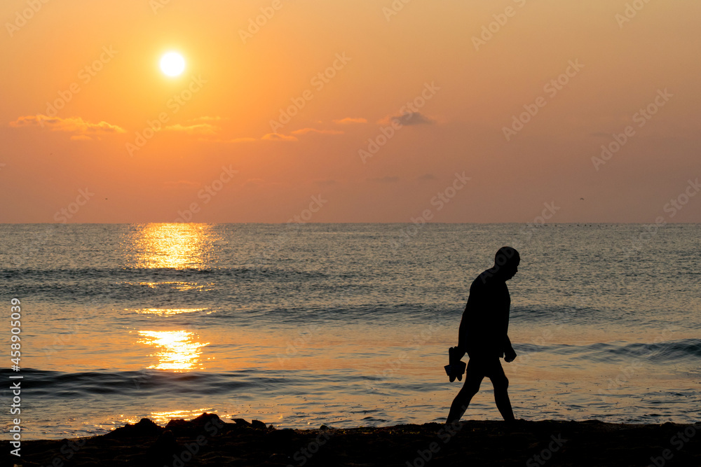 Seascape. Sunrise. A man (silhouette) with slippers in hand walks alone on the beach, thoughtful.