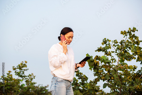 Female farmer entrepreneur using a digital tablet and smartphone to inspecting hazelnut orchard farm. Quality control, examining hazelnut tree plant for agriculture crop or food production industry