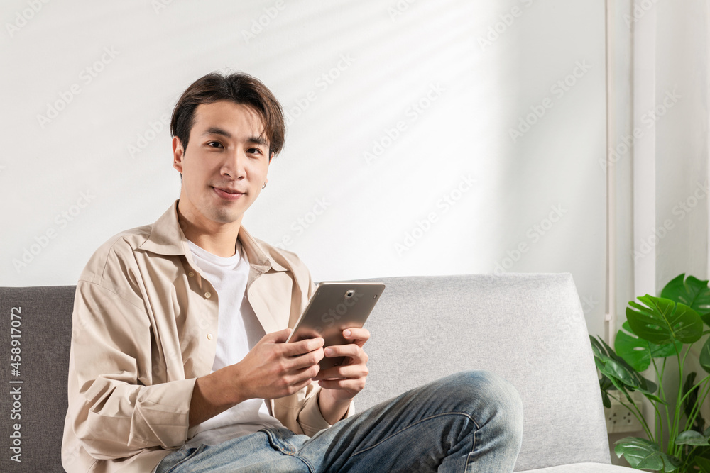 Happy smiling Asian student sits and relaxes on a couch using modern tablet browsing unlimited wireless internet, positive young man freelancer working uses wifi at a home apartment, technology idea Happy smiling Asian student sits and relaxes on a couch using modern tablet browsing unlimited wireless internet, positive young man freelancer working uses wifi at a home apartment, technology idea