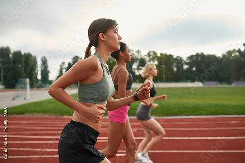 Side view of group of three professional young female runners running together on track field at the stadium
