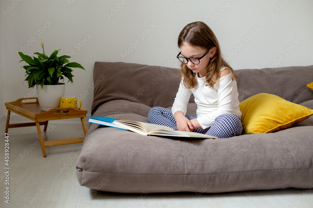 Obraz premium a cute little girl in glasses, jeans and a white turtleneck is reading a book, doing homework, preparing for exams sitting on the bed. home education. space for text. High quality photo