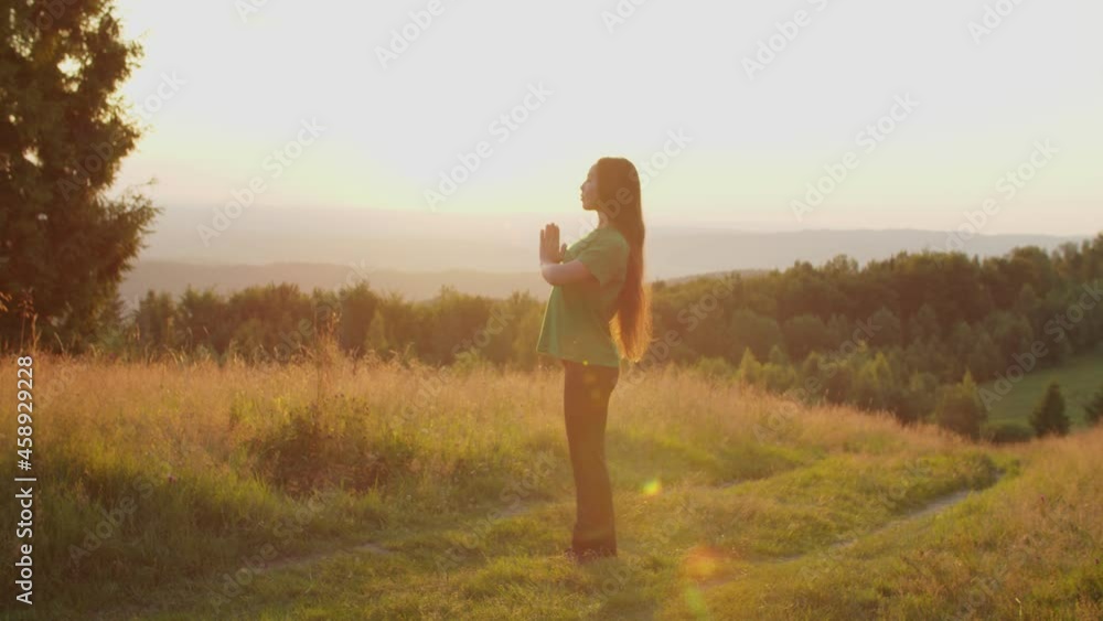 Portrait of serene lovely asian woman with eyes closed clasping hands in namaste gesture, meditating in prayer yoga pose while standing on mountain peak in rays of beautiful summer sunrise. Portrait of serene lovely asian woman with eyes closed clasping hands in namaste gesture, meditating in prayer yoga pose while standing on mountain peak in rays of beautiful summer sunrise.