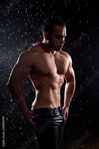 Handsome young caucasian man with naked torso posing at camera on black background. Drops of water spread about fitness body. The perfect figure on the background of water splashes. Portrait