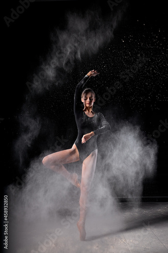 beautiful slender ballet dancer women wearing black bodysuit, sensually poses among flying flour which covers her body, on black studio background. Artistic, commercial, monochrome design.