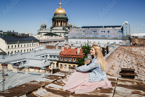 young happy woman tourist, in a flying pink skirt, sits on the rusty roof of a European city. view of the colonnade of the historic cathedral. roofs of St. Petersburg.