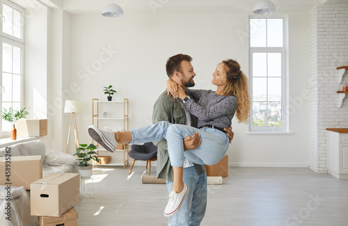Romantic portrait of joyful and satisfied young couple in love moving to their new home. Loving husband carried his smiling wife to their new bright and cozy living room. Moving concept.