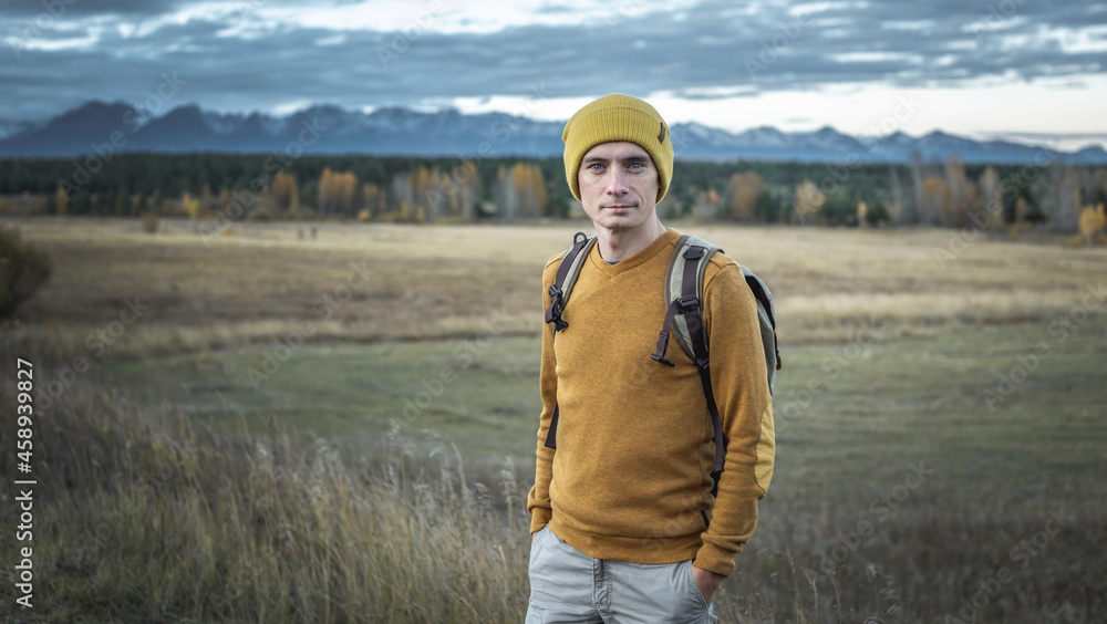 Man with a backpack in a yellow hat and a sweater in nature against the background of an autumn forest and mountains
