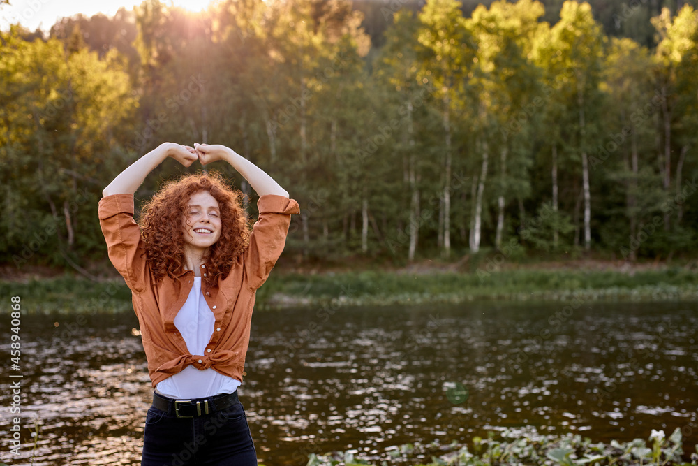 Fototapeta premium Portrait of cheerful happy redhead curly lady enjoying time in forest, raising hands up, smiling, caucasian lady is standing by lake, in the evening at sunset. in rural nature. people, beauty concept