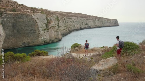 Hiking couple. Young People on Rocks Overlooking Nice View Over the Fomm ir-Riħ sea bay at Malta. Young couple with backpack walking to the edge of a cliff near the bank of the beautiful sea bay. photo