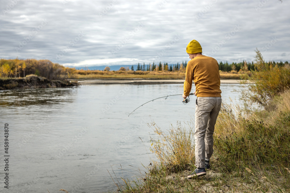Man in a yellow sweater and hat is fishing on the river bank in the autumn forest with a spinning rod in his hands