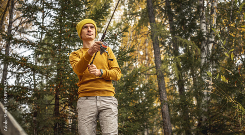 Man in a yellow sweater and hat is fishing on the river bank in the autumn forest with a spinning rod in his hands