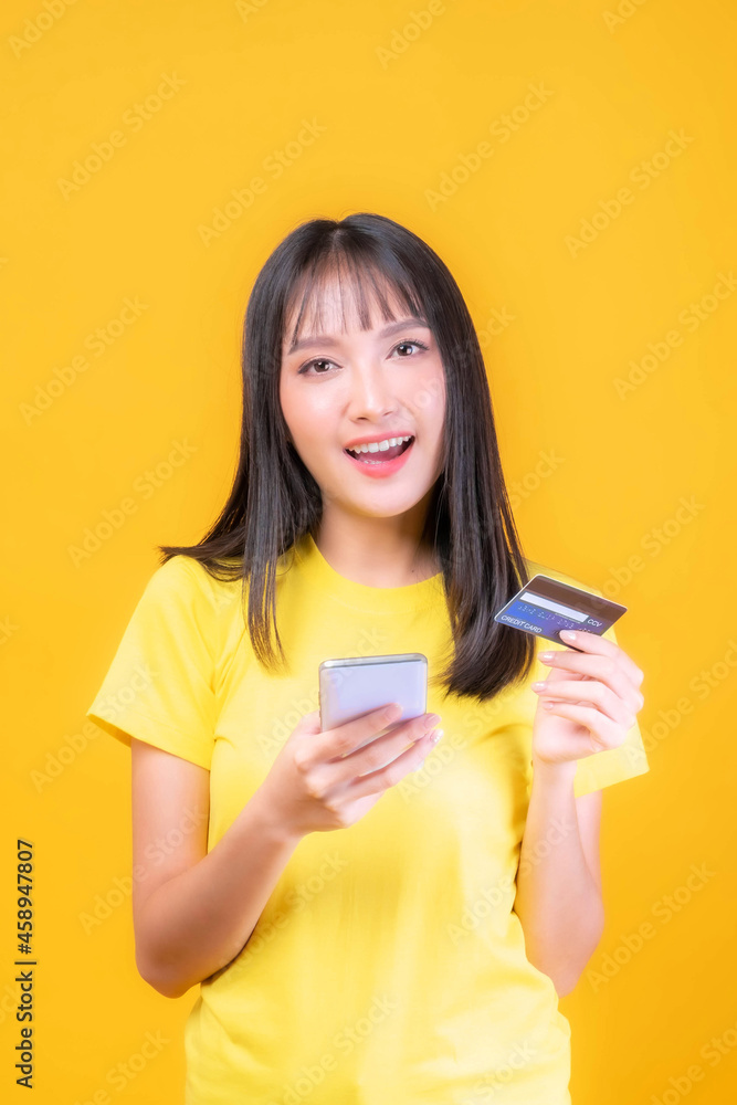 Naklejka premium beautiful Asian young woman cute girl with bangs hair style in yellow shirt holding credit card and smartphone ,mobile phone uses a online banking for pay online shopping isolated on yellow background