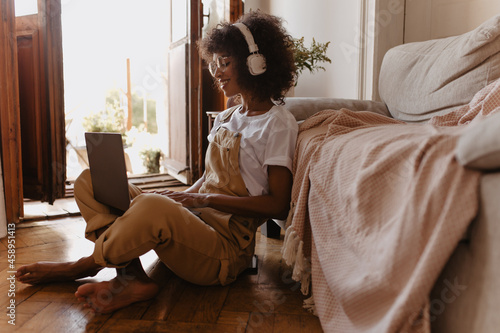 Photography side view of sophisticated African woman with legs crossed sitting on floor while working in laptop