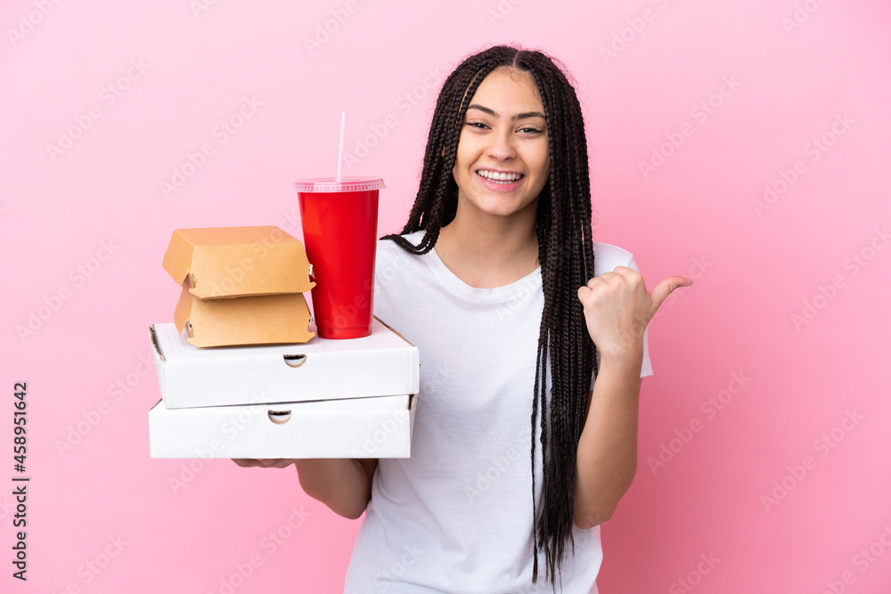 Teenager girl with braids holding pizzas and burgers over isolated pink background pointing to the side to present a product Teenager girl with braids holding pizzas and burgers over isolated pink background pointing to the side to present a product