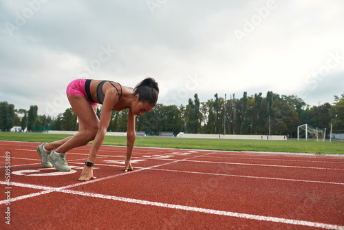 Professional young female runner in sportswear starting race on track field at stadium
