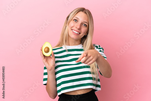 Young caucasian woman holding an avocado isolated on pink background pointing front with happy expression