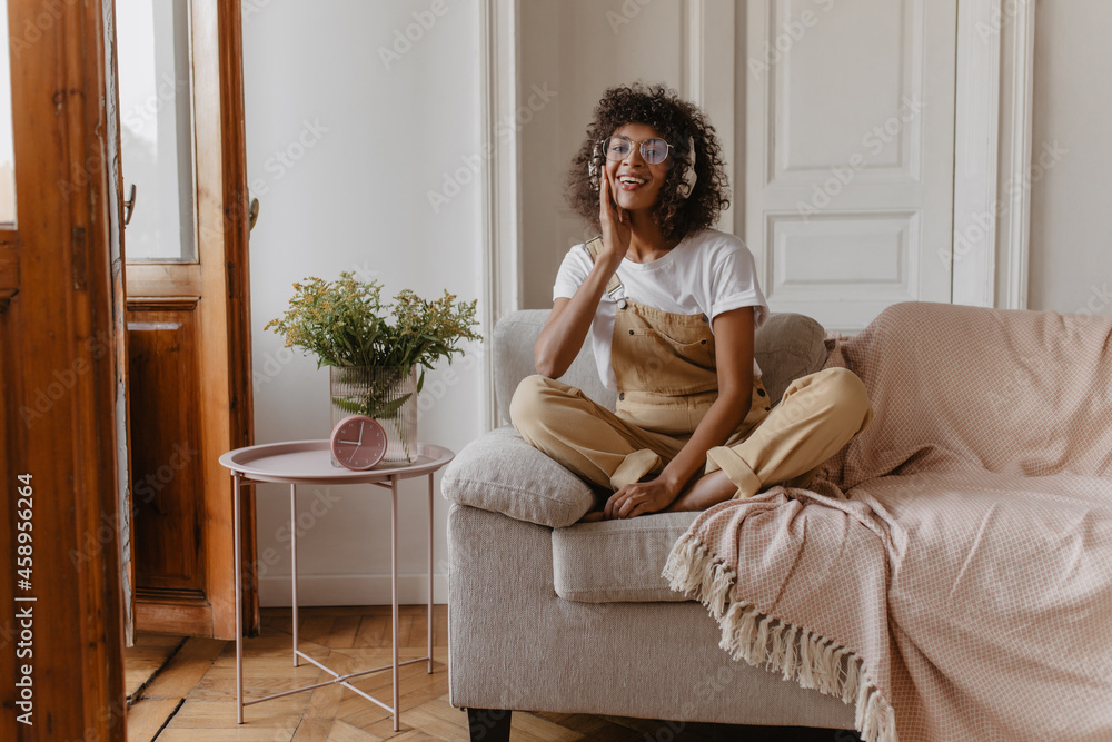 stunningly sweet girl sits cross-legged on white sofa, huddled in light room. swarthy girl in headphones, glasses is dressed in white T-shirt and jumpsuit on top. stunningly sweet girl sits cross-legged on white sofa, huddled in light room. swarthy girl in headphones, glasses is dressed in white T-shirt and jumpsuit on top.