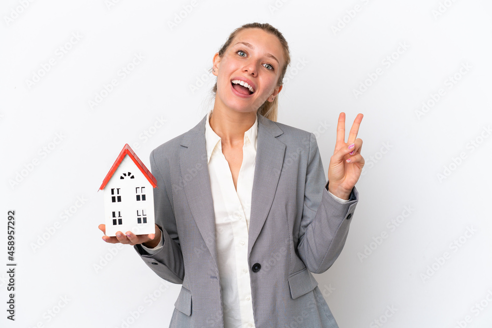Real estate agent woman holding a toy house isolated on white background smiling and showing victory sign Real estate agent woman holding a toy house isolated on white background smiling and showing victory sign
