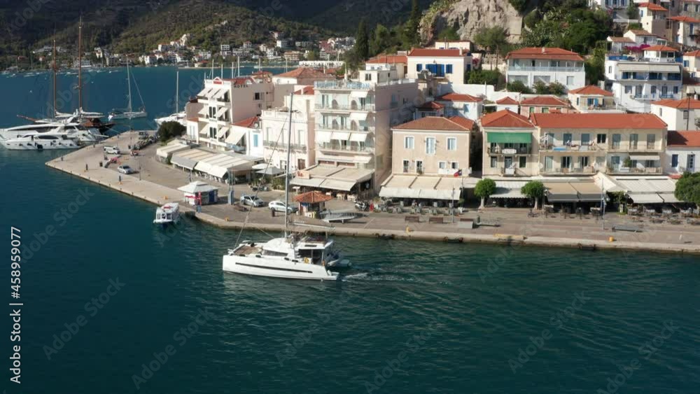 Catamaran Boat Sailing By The Terminal With Waterfront Houses In Poros Island, Greece. aerial Catamaran Boat Sailing By The Terminal With Waterfront Houses In Poros Island, Greece. aerial