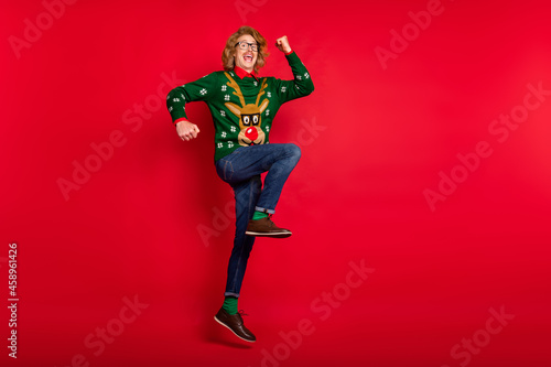 Photo of funny lucky young guy dressed print pullover smiling jumping rising arms empty space isolated red color background