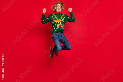 Photo of handsome funky young guy dressed print pullover jumping rising fists smiling isolated red color background