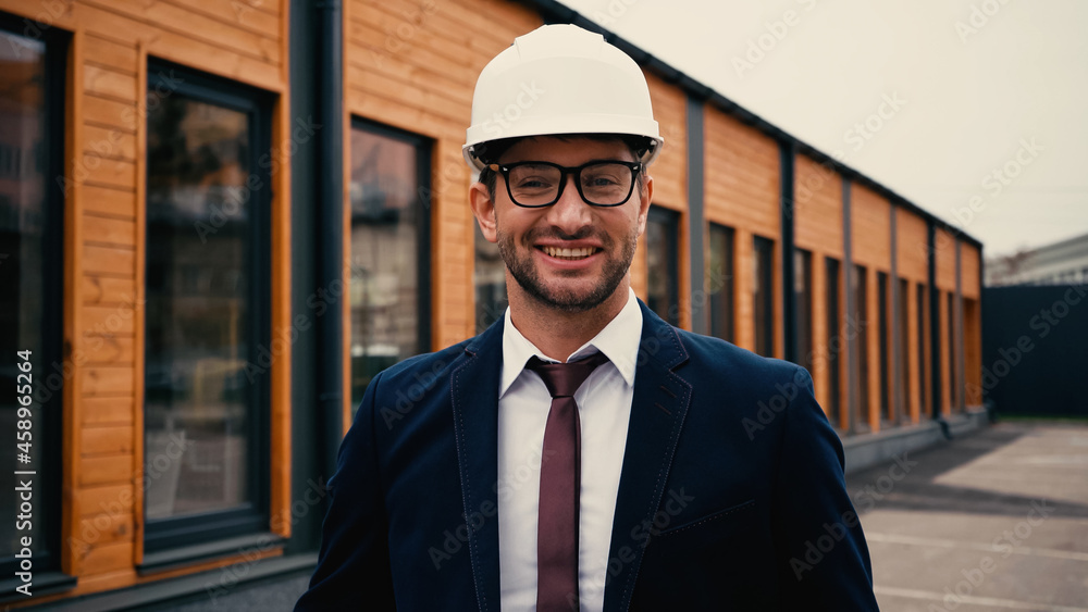 Fototapeta premium Smiling engineer in white hardhat looking at camera near building outdoors