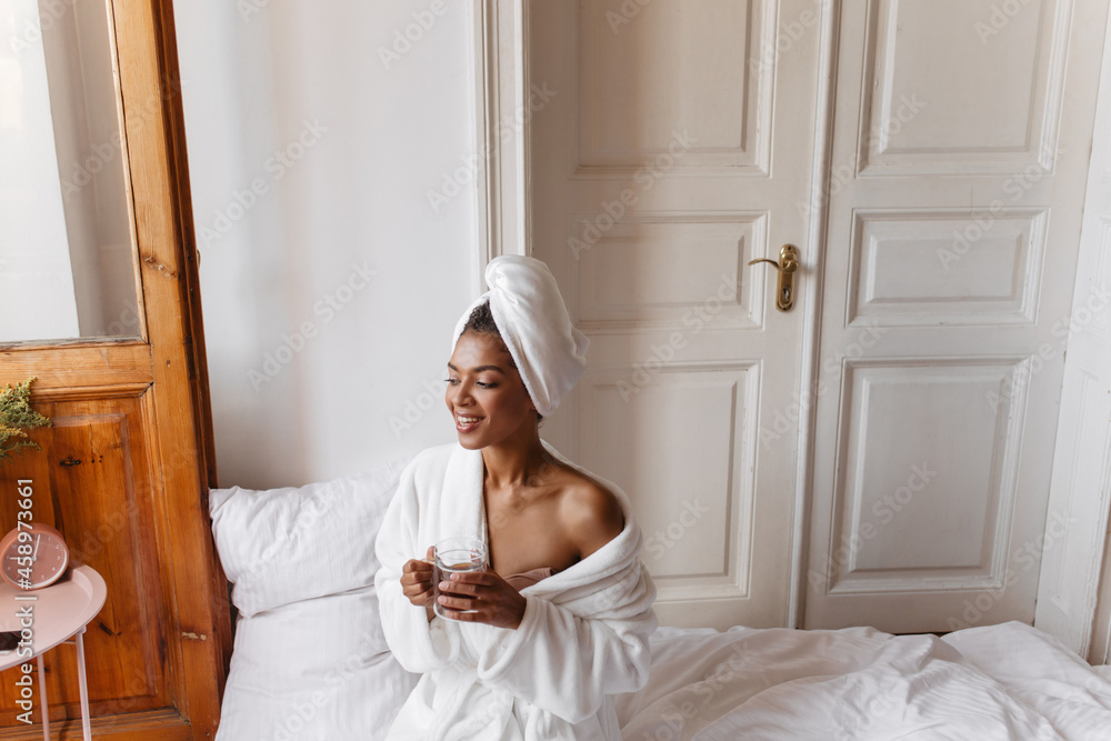 top view of gorgeous young African woman in early morning sitting on bed in bright room. after shower, madame drinks aromatic coffee in white robe with towel on her head. top view of gorgeous young African woman in early morning sitting on bed in bright room. after shower, madame drinks aromatic coffee in white robe with towel on her head.