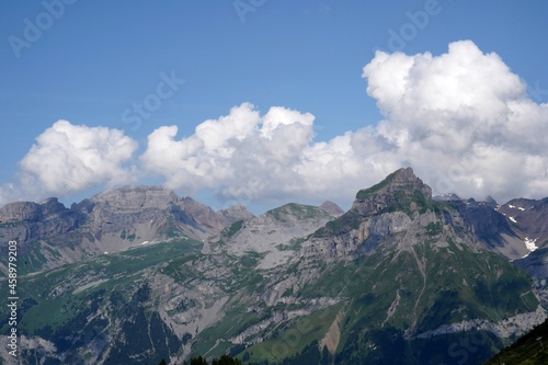 Mountains surrounding village Engelberg in Switzerland in summertime. Alpine meadows in mountains are dark green. There are thick white clouds coming from behind the mountains.  © Lucia