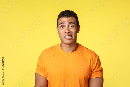 Yikes, awkward. Embarrassed young handsome man caught on lie, feel indesicive and slightly worried, cringe, smiling nervously and look camera with reluctance, stand yellow background photo