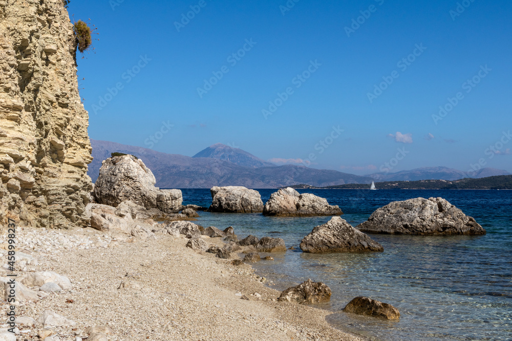 White pebbles and rocks cliffs beach with azure clear water and mountains in distance on coast of Lefkada island in Greece. Summer wild nature vacation travel to Ionian Sea