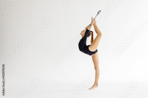 slim artistic teenager girl in black leotard trains on white background with clubs in her hands in rhythmic gymnastic exercise, children's professional sports