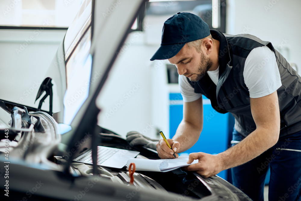 Auto mechanic writes data while examining engine at repair shop. Auto mechanic writes data while examining engine at repair shop.