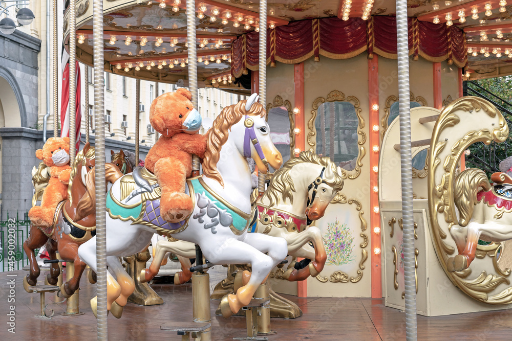 Teddy bear in a medical mask rides on a children's circular carousel.