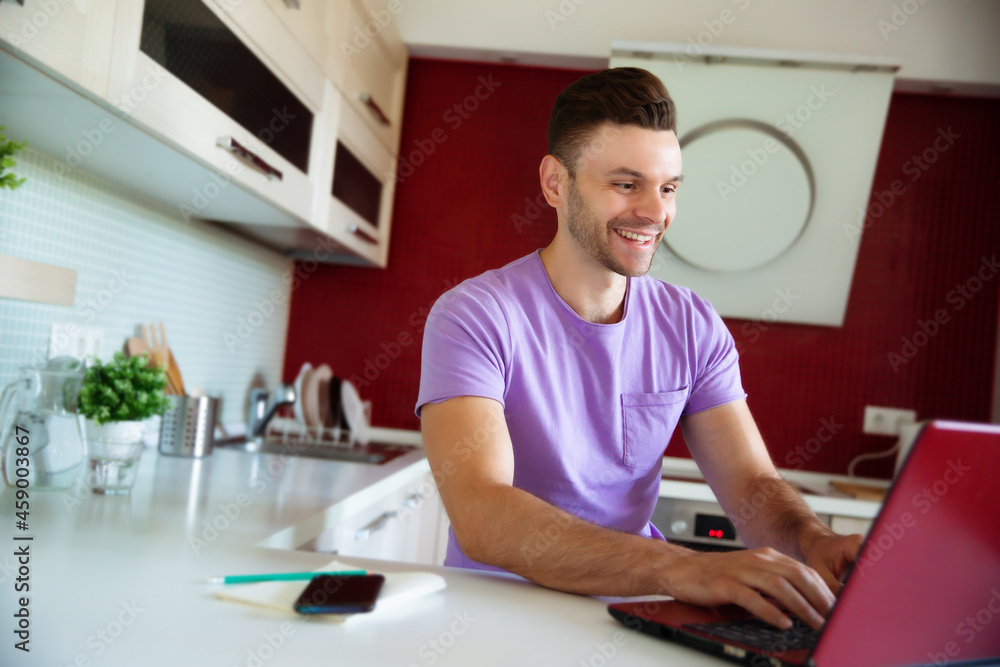 Happy handsome smiling young man in casual clothes is sitting while working on his laptop in the modern kitchen at home Happy handsome smiling young man in casual clothes is sitting while working on his laptop in the modern kitchen at home