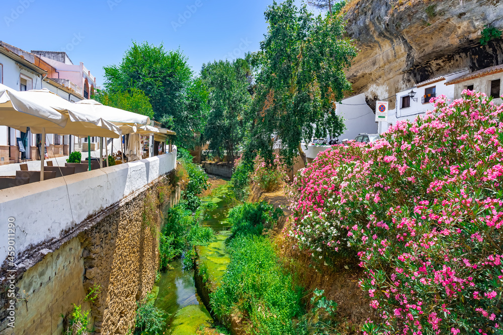 Cauce de un río con exuberante vegetación a su paso por el centro de un pueblo de calles blancas desde Setenil de las Bodegas, provincia de Cádiz, Andalucía, España.