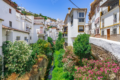 Cauce de un río con exuberante vegetación a su paso por el centro de un pueblo de calles blancas desde Setenil de las Bodegas, provincia de Cádiz, Andalucía, España.