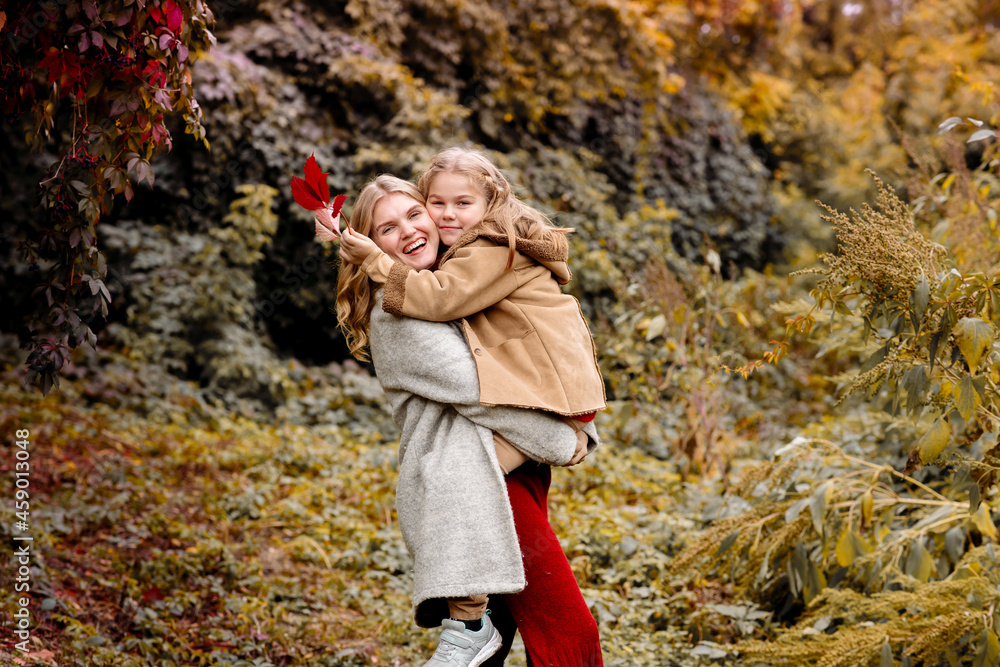 A young adult blonde mother with a 7-year-old daughter on a walk in the autumn park hug and laugh

