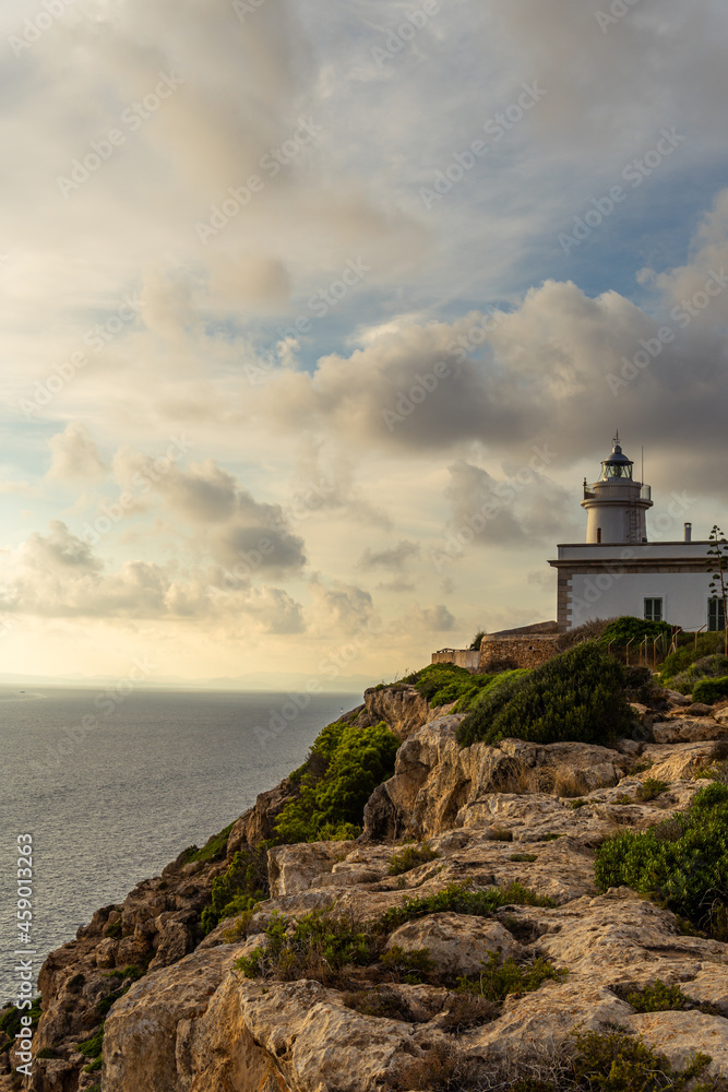 Cap Blanc lighthouse on the rocky coast of the island of Mallorca. In the background s seascape of the Mediterranean Sea at sunset with clouds. Sublime landscape