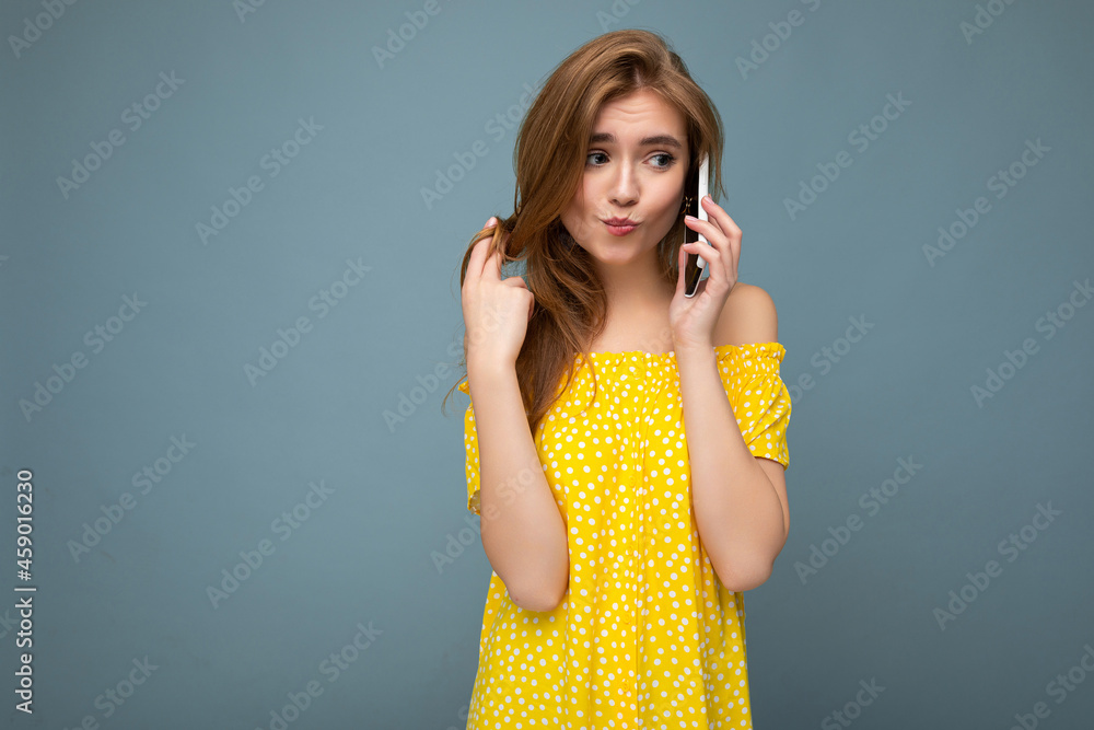 Sexy attractive positive smiling young blonde woman wearing stylish yellow summer dress standing isolated over blue background holding and talking on mobile phone looking to the side Sexy attractive positive smiling young blonde woman wearing stylish yellow summer dress standing isolated over blue background holding and talking on mobile phone looking to the side