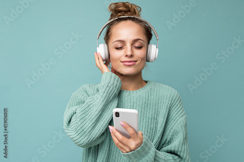Photo shot of beautiful joyful smiling young female person wearing stylish casual outfit isolated over colorful background wall wearing white bluetooth wireless earphones and listening to music and photo