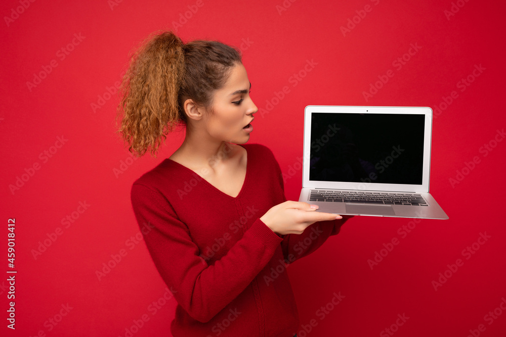 Beautiful charming fascinating amazed young curly dark blond woman wearing red sweater standing isolated over red wall background holding computer laptop with empty copy space mock up pointing to the Beautiful charming fascinating amazed young curly dark blond woman wearing red sweater standing isolated over red wall background holding computer laptop with empty copy space mock up pointing to the