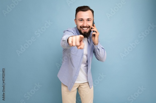 Photo shot of positive smiling good looking young brunet bearded man wearing casual blue shirt and white t-shirt poising isolated on blue background with empty space holding in hand and communicating