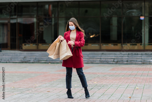 Portrait of young fashion woman wearing protective mask with shopping bags on city street