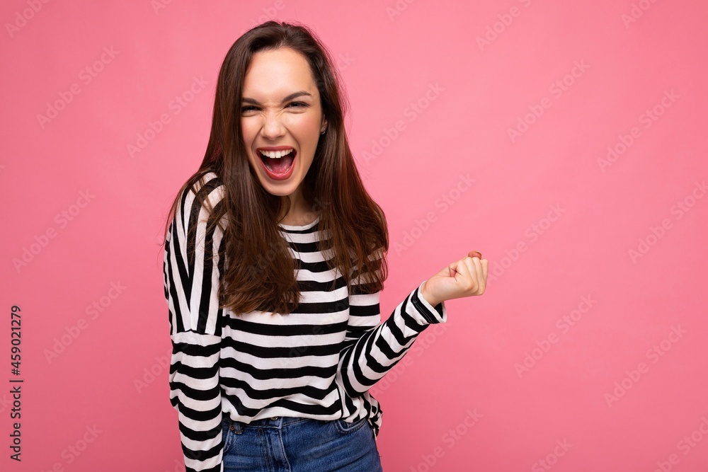 Portrait of young emotional positive happy attractive brunette woman with sincere emotions in casual striped pullover isolated on pink background with free space and celebrating winning shouting yes Portrait of young emotional positive happy attractive brunette woman with sincere emotions in casual striped pullover isolated on pink background with free space and celebrating winning shouting yes
