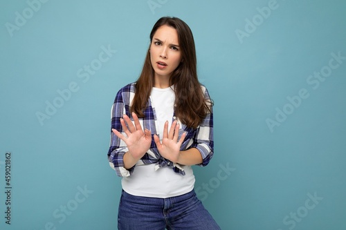 Young serious dissatisfied beautiful brunette woman with sincere emotions wearing trendy check shirt standing isolated on blue background with empty space and showing stop gesture saying no. Negative photo