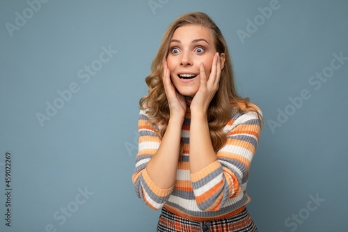 Photo of young surprised pretty nice blonde curly woman with sincere emotions wearing casual striped pullover isolated on blue background with free space